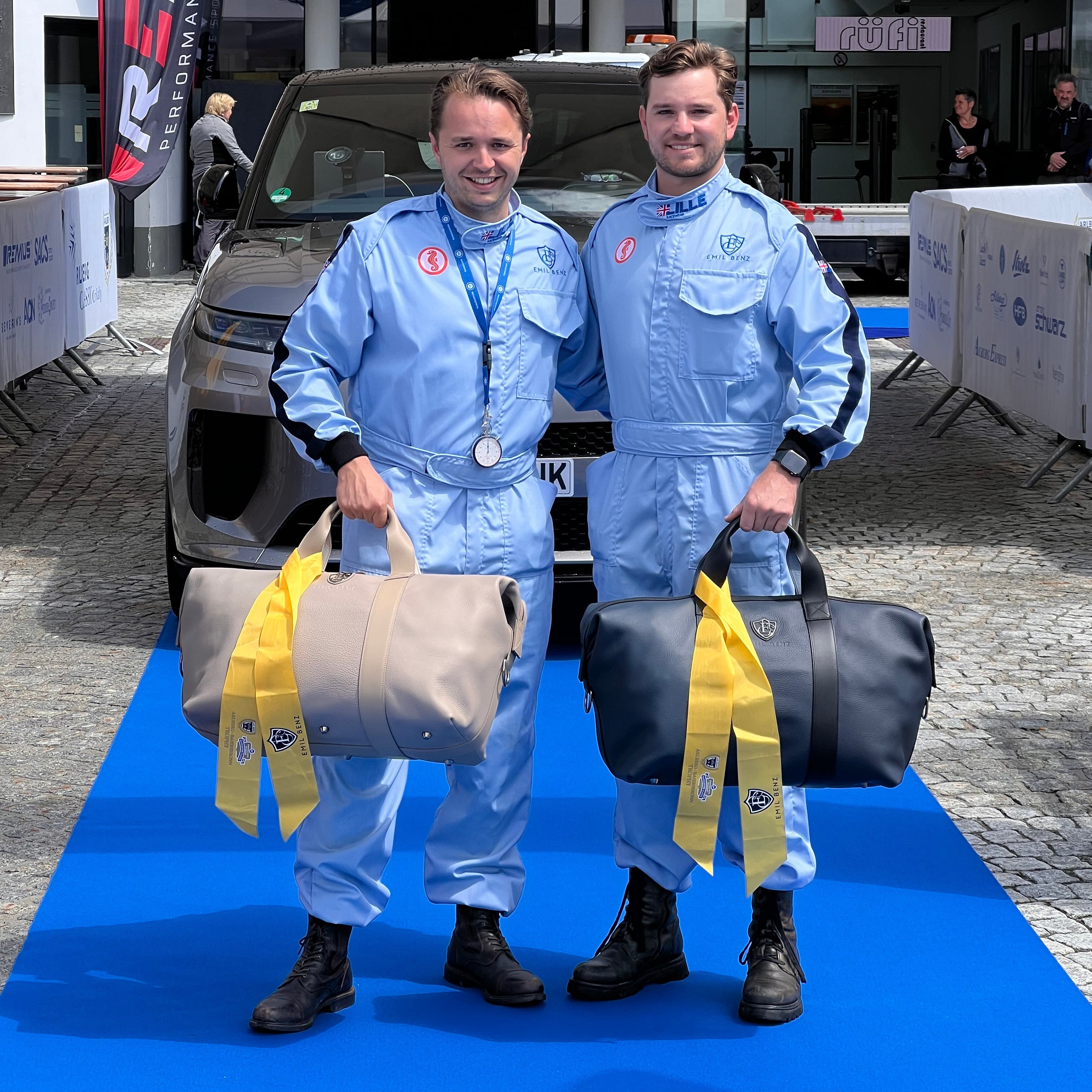 Two individuals in blue racing suits holding duffel bags on a blue mat with a car and event setup in the background.